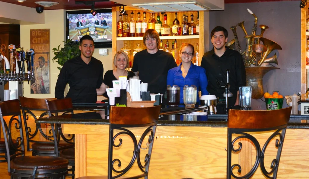 Lively bar interior with five smiling staff, jazz sign, and instrument sculpture.