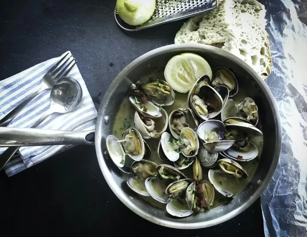 Overhead: Pan of cooked clams, lemon, rustic bread, and cutlery.