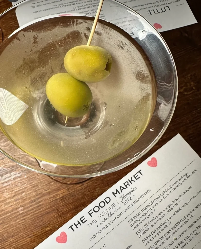 Martini with two olives on wooden table beside restaurant menu.
