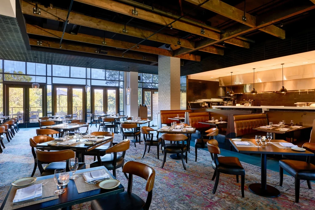 Modern restaurant dining room with set tables, outdoor view, and open kitchen.