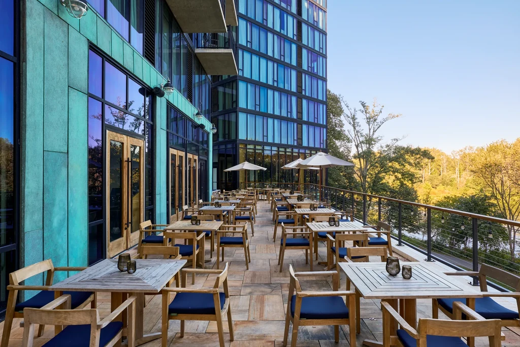Empty outdoor patio with blue chairs, tables, umbrellas, and modern buildings.