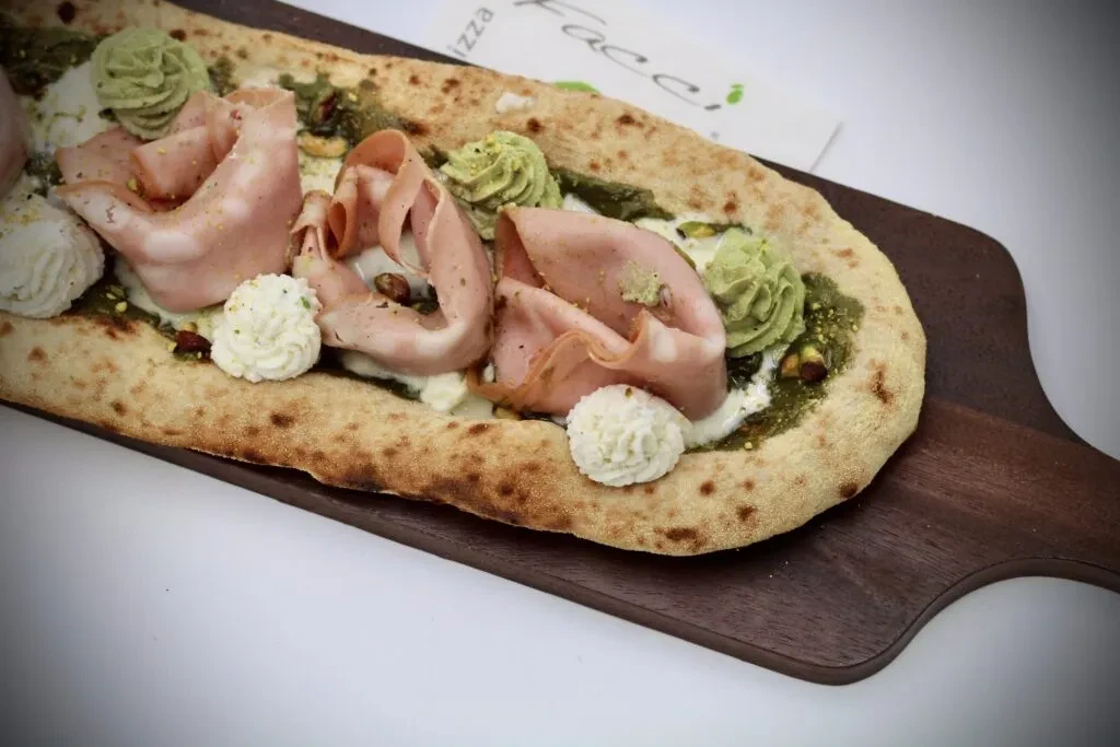 Elongated flatbread with mortadella, white cheese, green pesto, and pistachios.