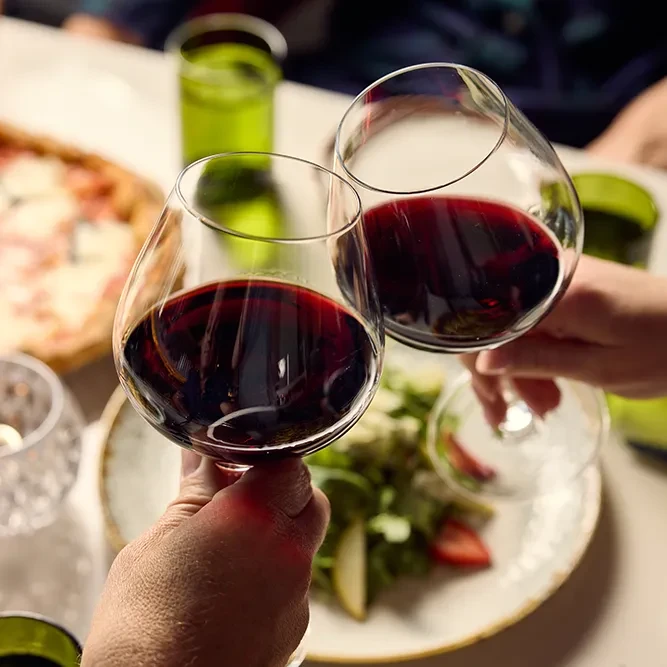 Hands clinking red wine glasses at a warm social dinner.