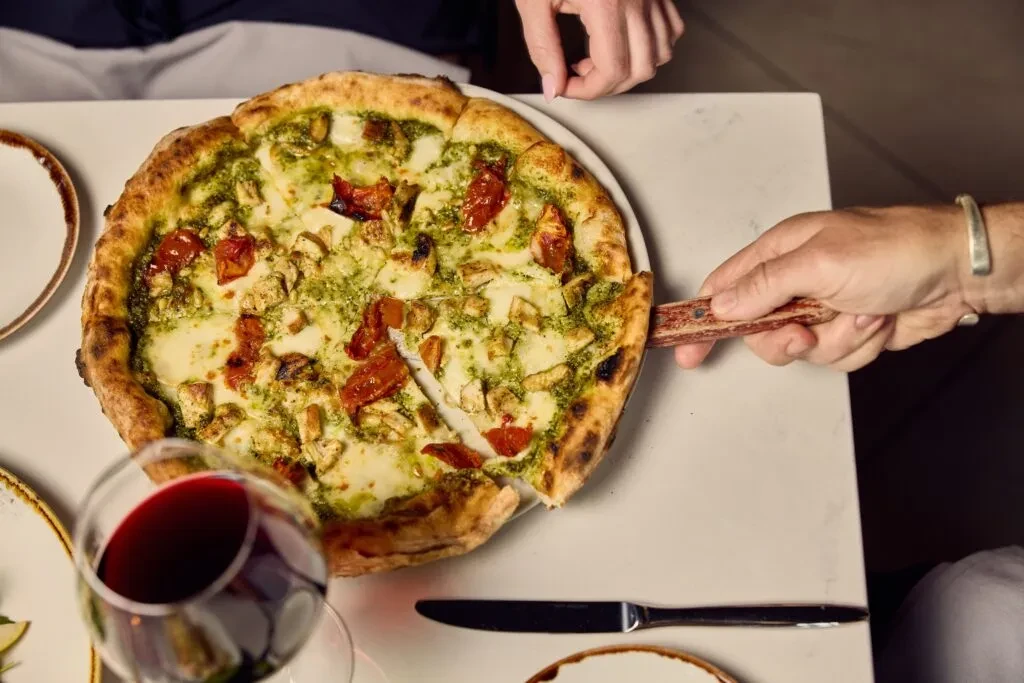 Overhead view: hand lifts pesto chicken pizza slice with red toppings.