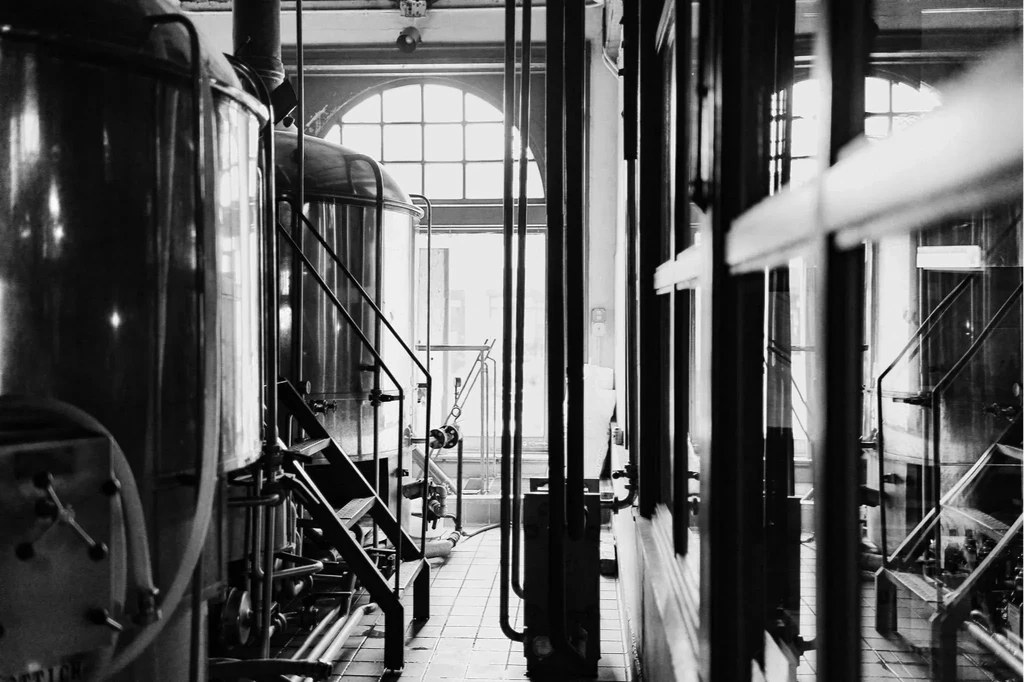 Black and white brewery interior with large tanks, pipes, and arched windows.