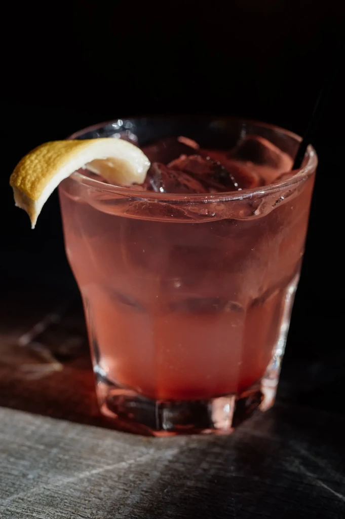 Reddish-pink cocktail in faceted glass with ice and twisted lemon peel.