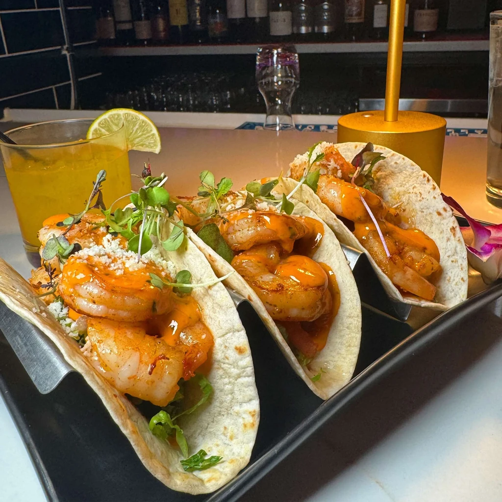 Three shrimp tacos with microgreens, yellow cocktail, blurred bar setting.