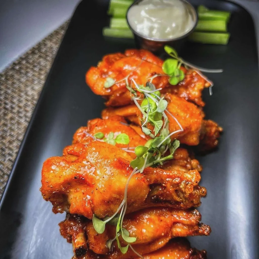 Chicken wings with shiny orange sauce, green microgreens, dip, and celery.