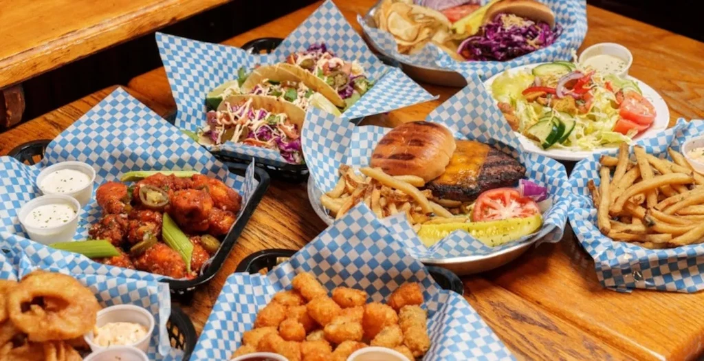 Rustic table filled with casual dishes: wings, tacos, burgers, fries, salad.