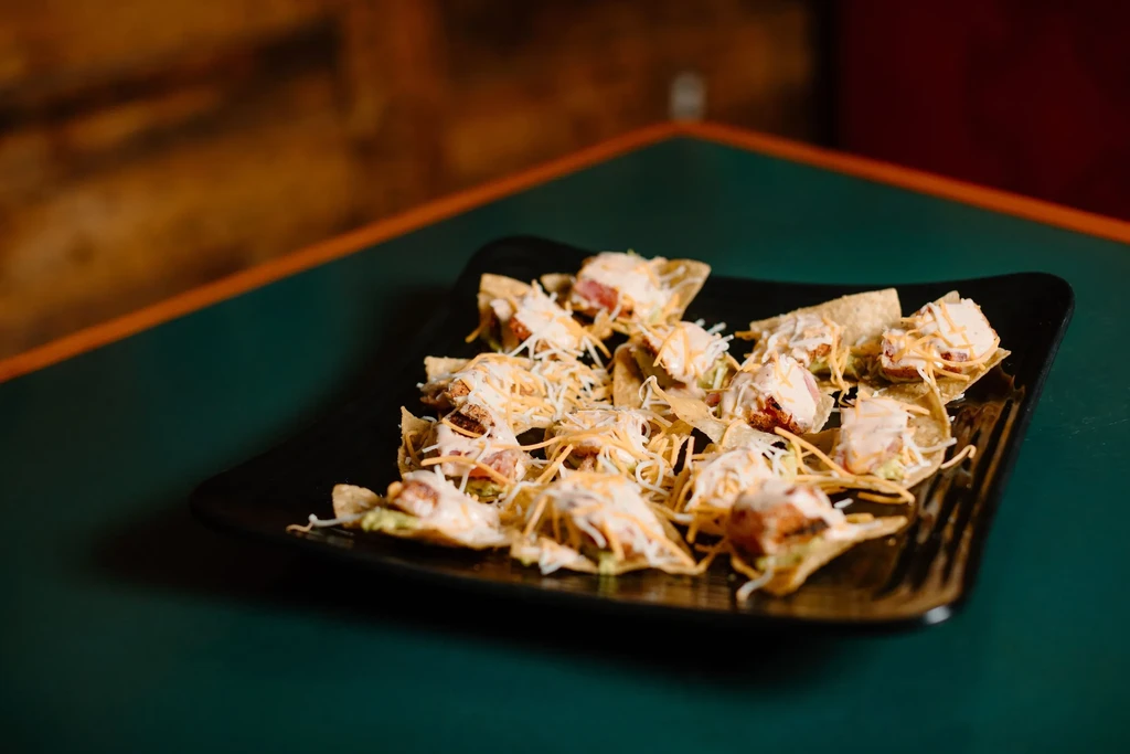 Black plate of mini-nachos topped with chicken, guacamole, cheese, and sauce.