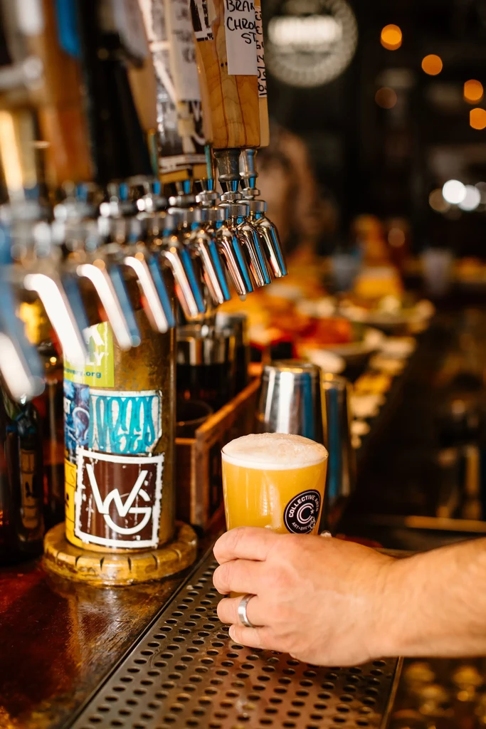 Hand holding frothy amber beer glass on bar drip tray.