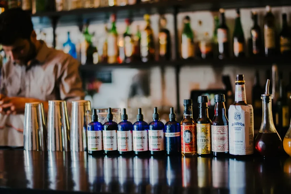 Bitters bottles and shakers on a bar counter with a blurred bartender.
