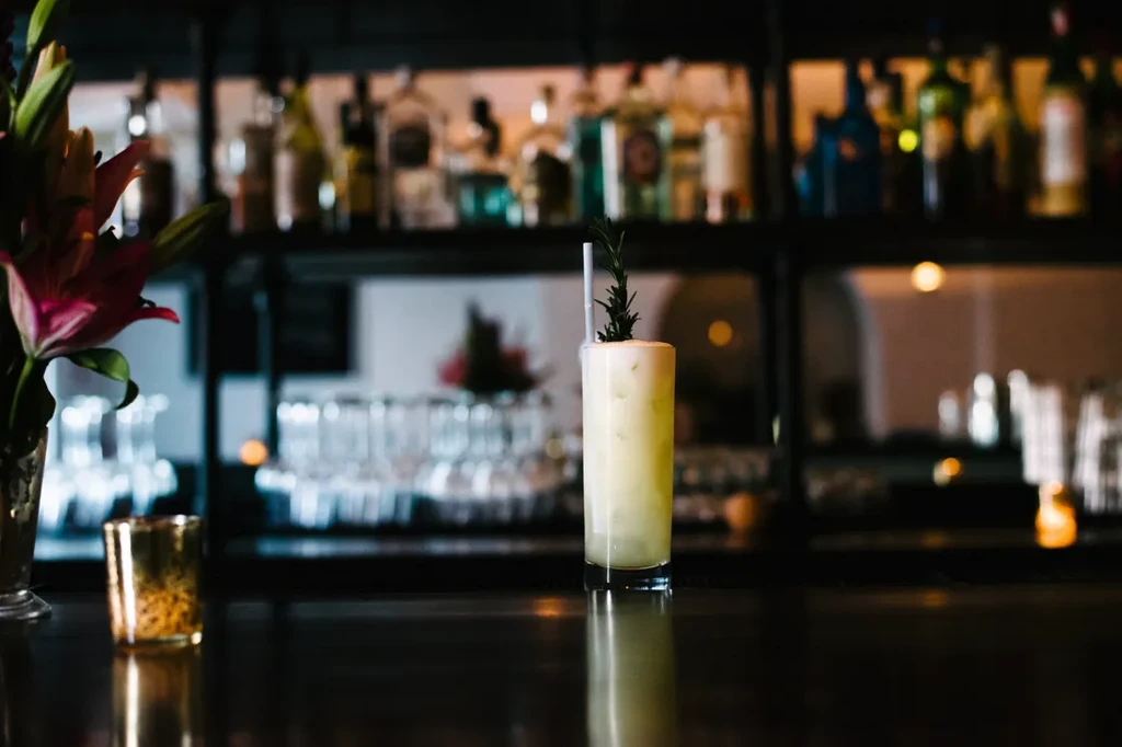 Cocktail with frothy top, rosemary, straw on bar, blurred liquor bottles.