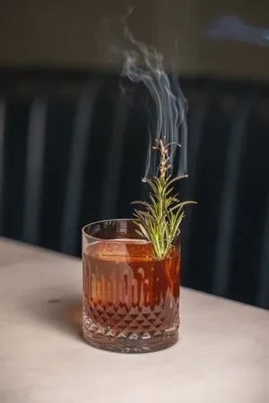 Amber cocktail in cut glass with smoking rosemary garnish.