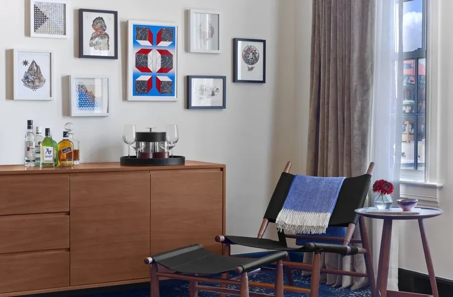 Wooden sideboard bar setup under gallery wall, with modern chair and flowers.