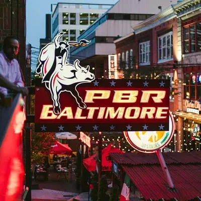 Nighttime urban street with an illuminated PBR Baltimore sign and a man.