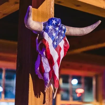 American flag-painted bull skull hanging on a rustic wooden beam.
