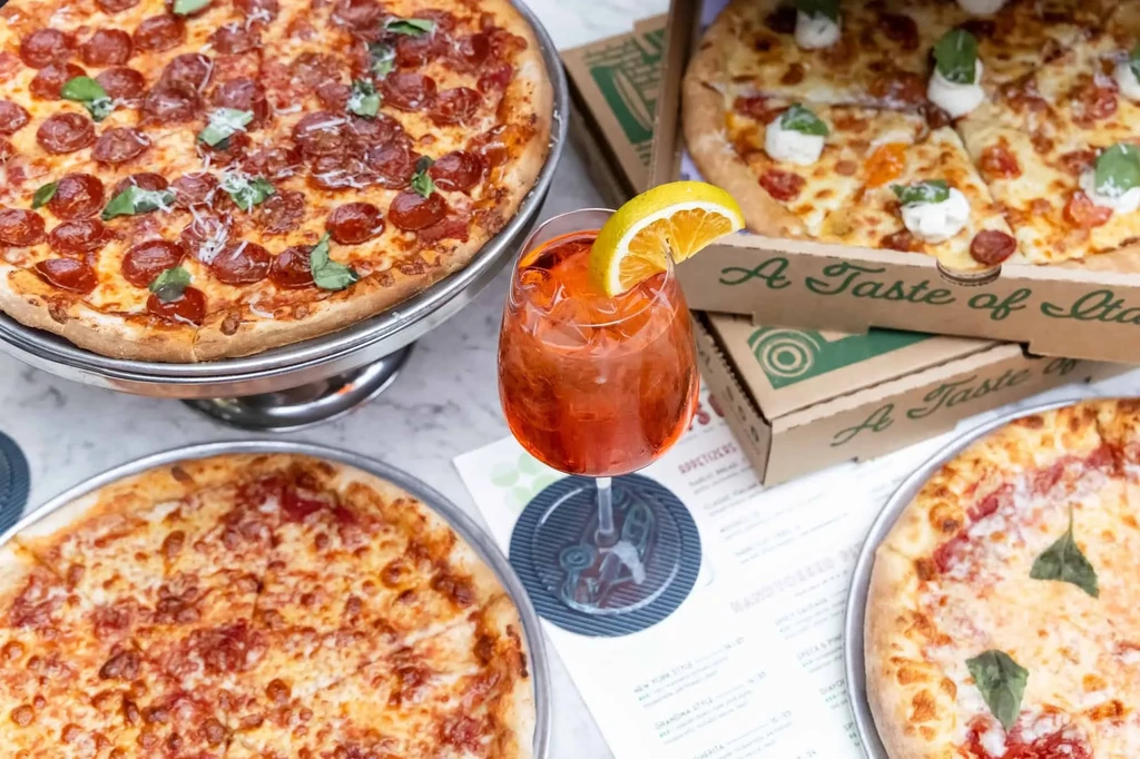 Overhead shot of four pizzas and an orange cocktail on marble.