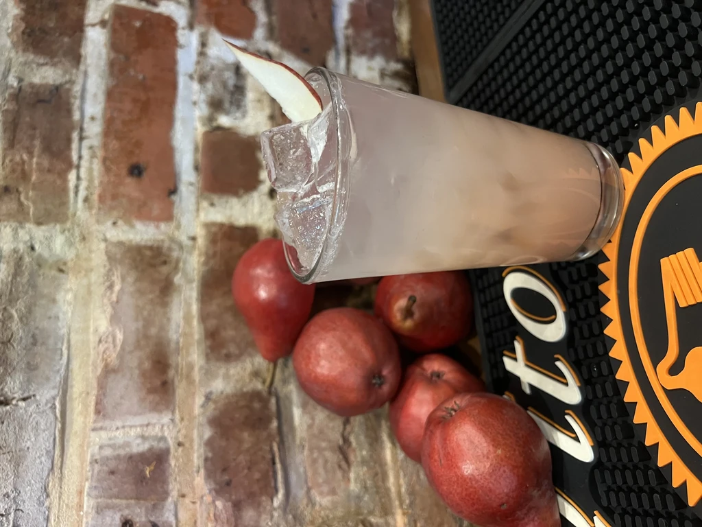 Cloudy cocktail with pear slice, ice, and red pears on bar.