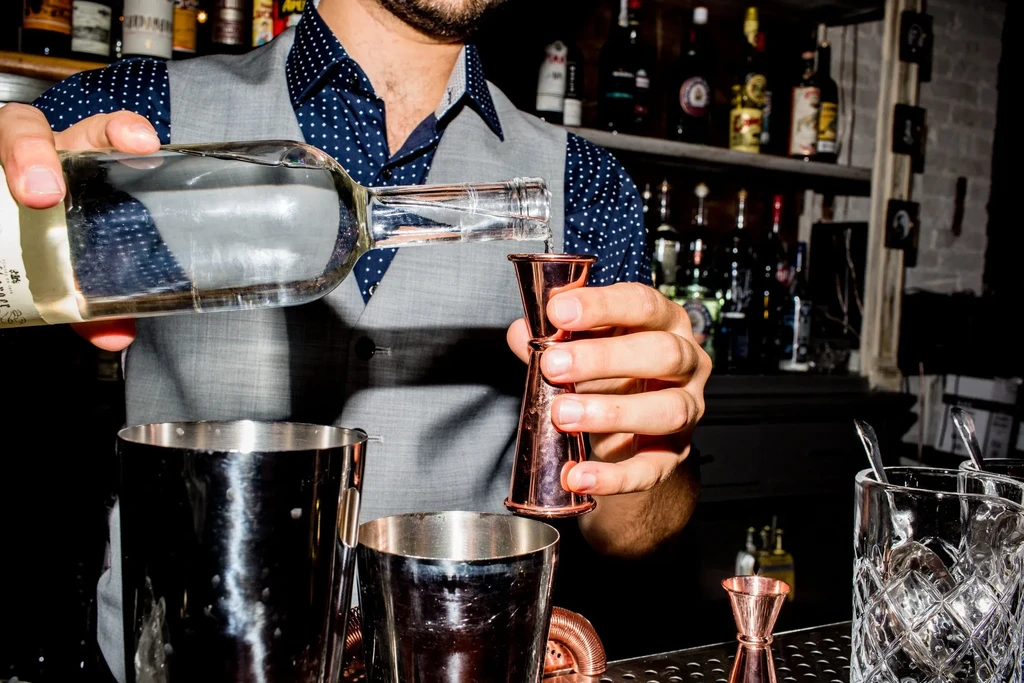 Bartender's hands pour spirits into a copper jigger at a dim bar.