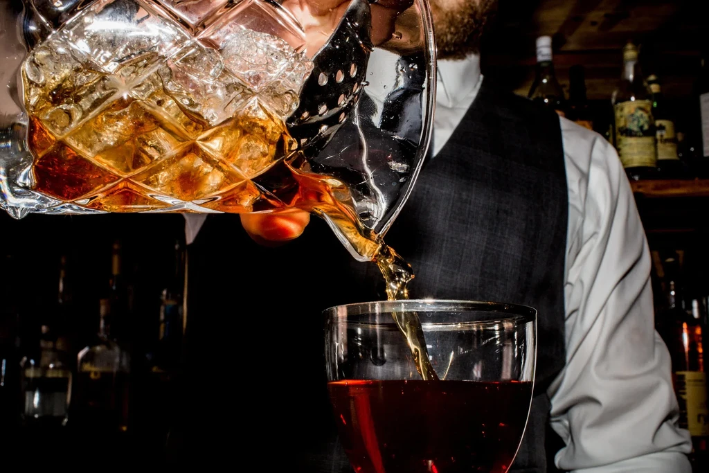 Bartender in vest pours amber liquid from ornate crystal mixing glass.