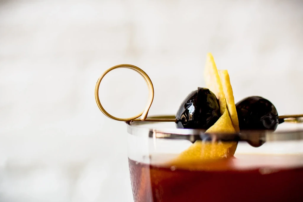 Two dark olives and yellow citrus peel on a golden cocktail pick.