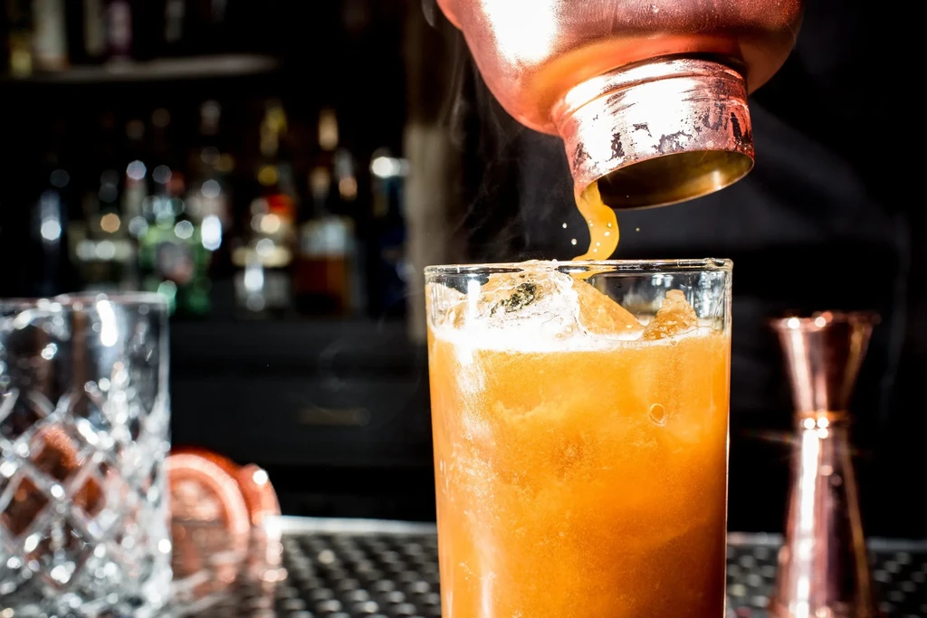 Copper shaker pouring liquid into frosty orange drink with ice.