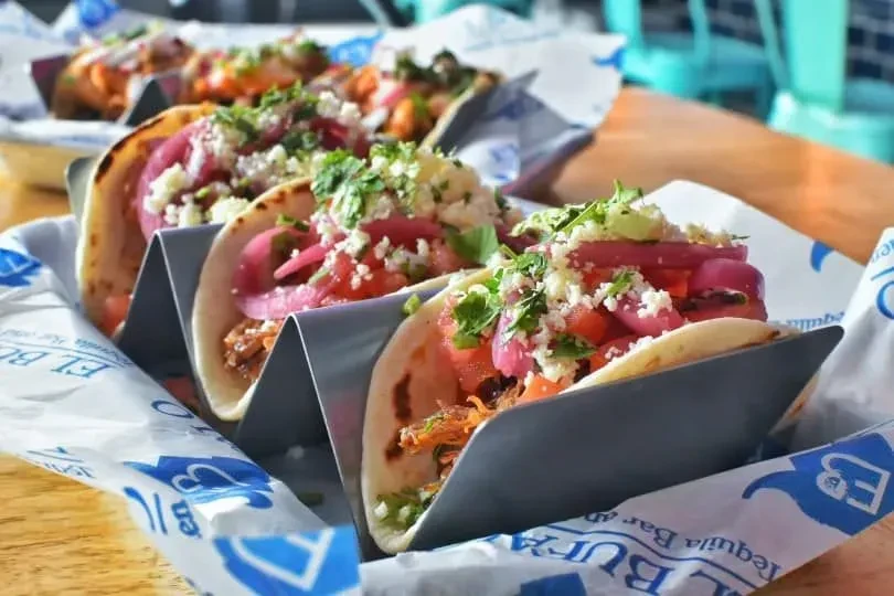 Three tacos bursting with colorful fillings in a branded basket.