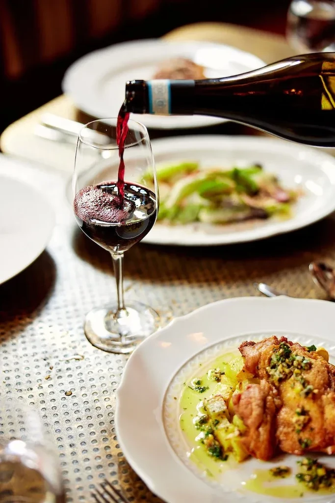 Red wine pours into a glass beside pan-seared meat with pesto.