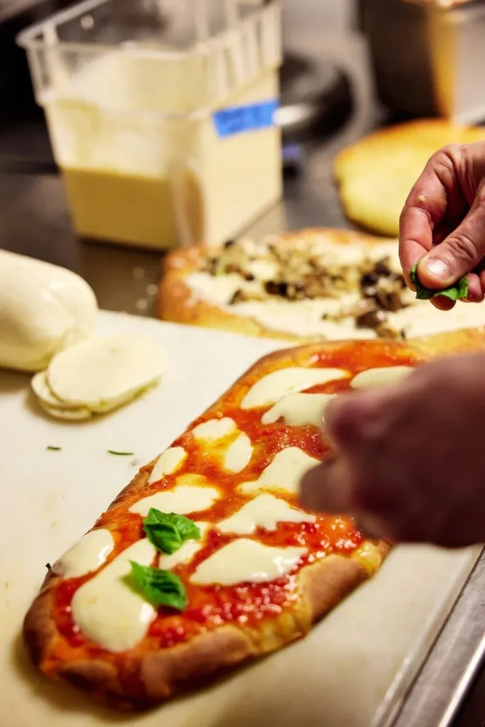 Chef's hands place basil on a rectangular pizza with tomato and mozzarella.