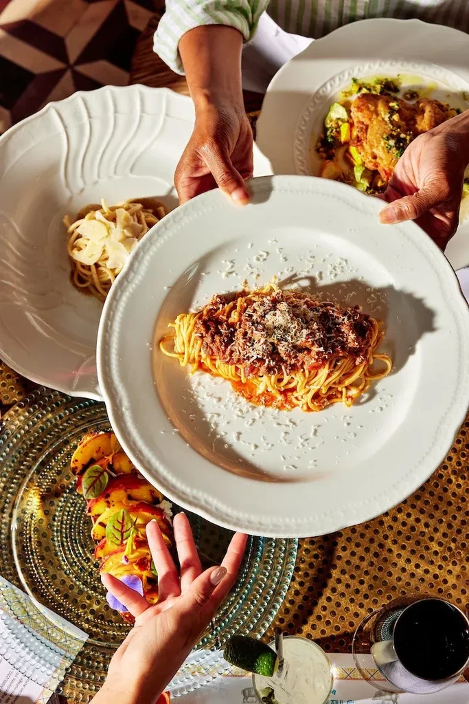 Overhead view of hands serving spaghetti bolognese and multiple upscale dishes.