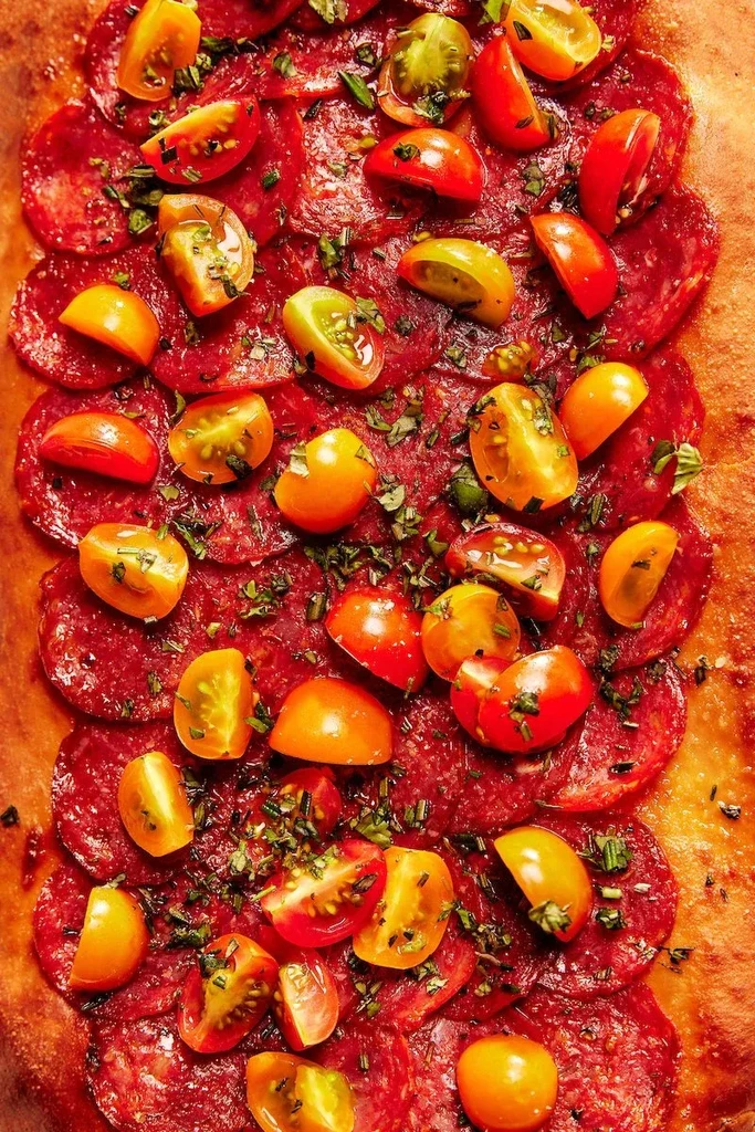 Close-up of pizza with cured meat, cherry tomatoes, and green herbs.