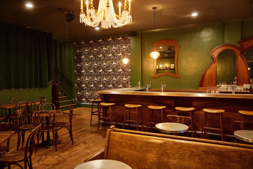 Dimly lit vintage bar interior with dark wood, green walls, and tables.