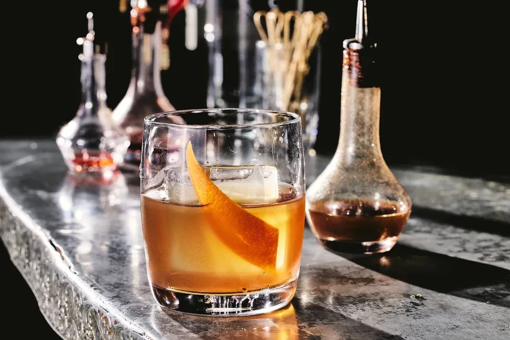 Amber cocktail with ice and orange peel on gleaming metal bar.