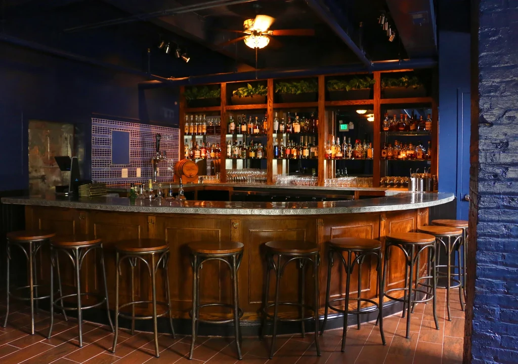 Curved wooden bar counter, eight barstools, and shelves of bottles.