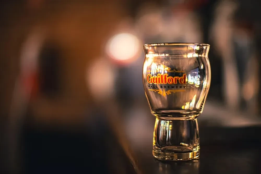 Empty glass with "Guilford Hall Brewery" logo on dark reflective surface.