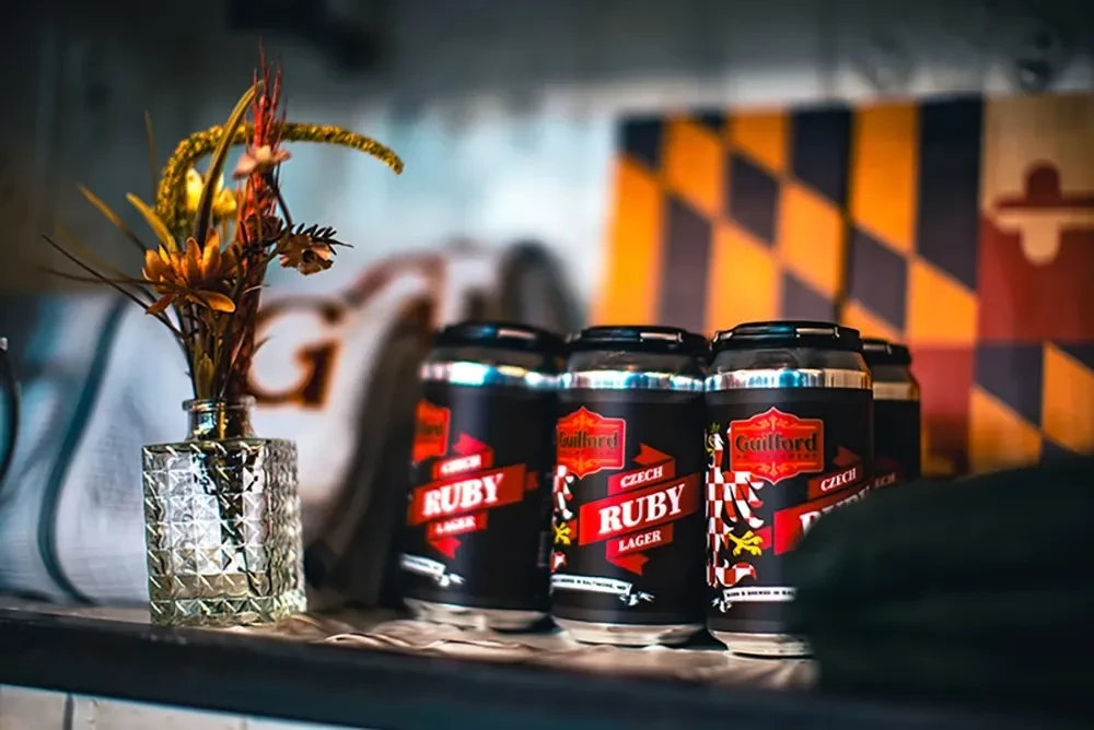 Black 'Guilford Czech Ruby Lager' cans, flowers, and a Maryland flag.