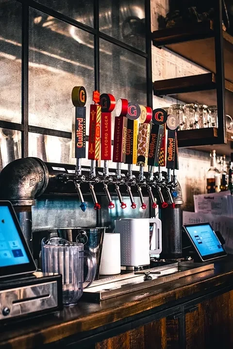 Eight branded beer taps on a rustic pipe fixture at a dark bar.