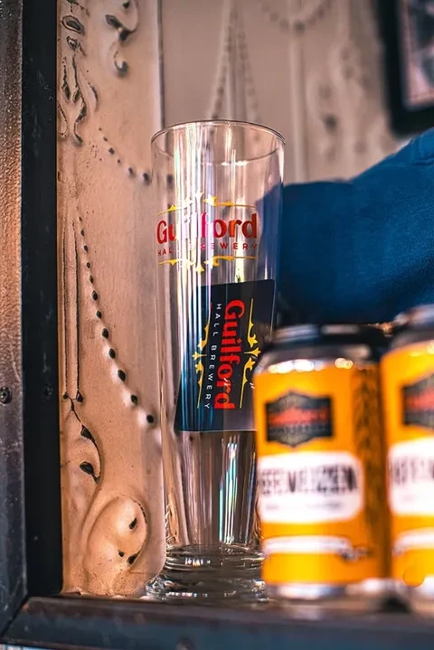 Empty Guilford Hall Brewery glass, textured wall, stacked beer cans, warm light.
