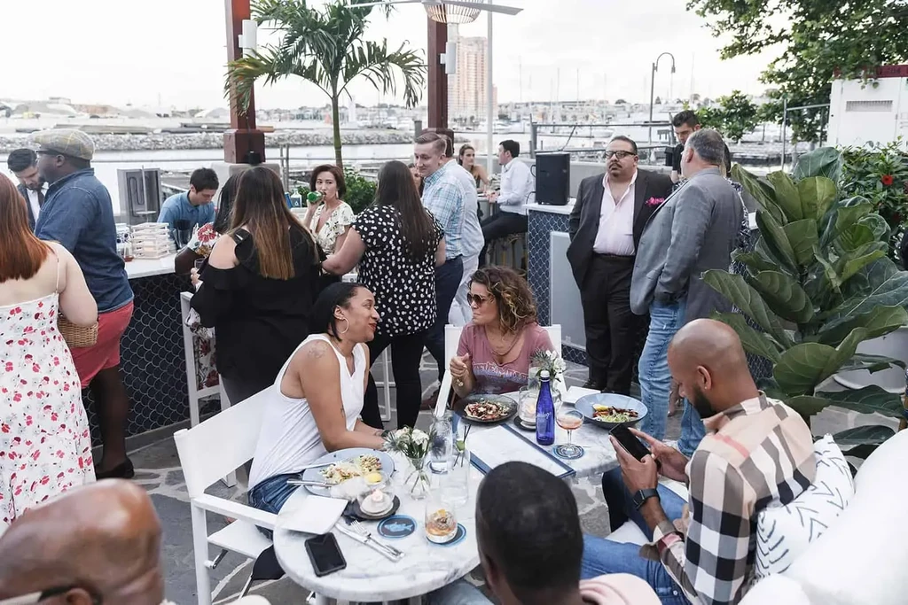 Lively outdoor bar/restaurant overlooking water, patrons dining and socializing.