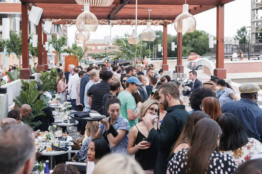Crowded outdoor restaurant patio with pergola, patrons, and marble-top tables.