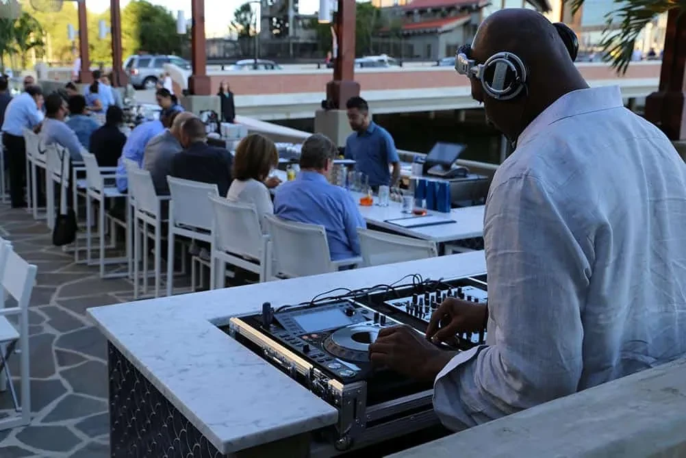 DJ from behind with headphones at lively outdoor bar, patrons.