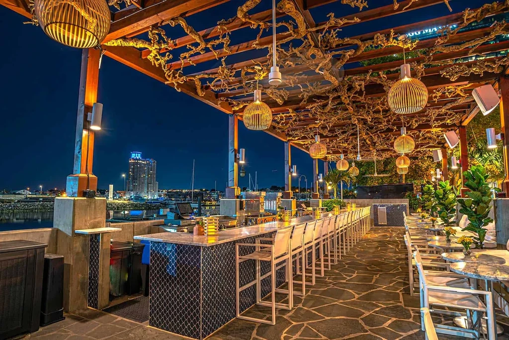 Illuminated outdoor patio bar with pergola, waterfront, and city skyline.