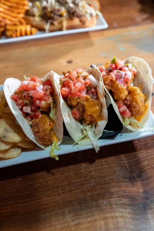 Three tacos with fried protein, red salsa, creamy sauce, and chips.