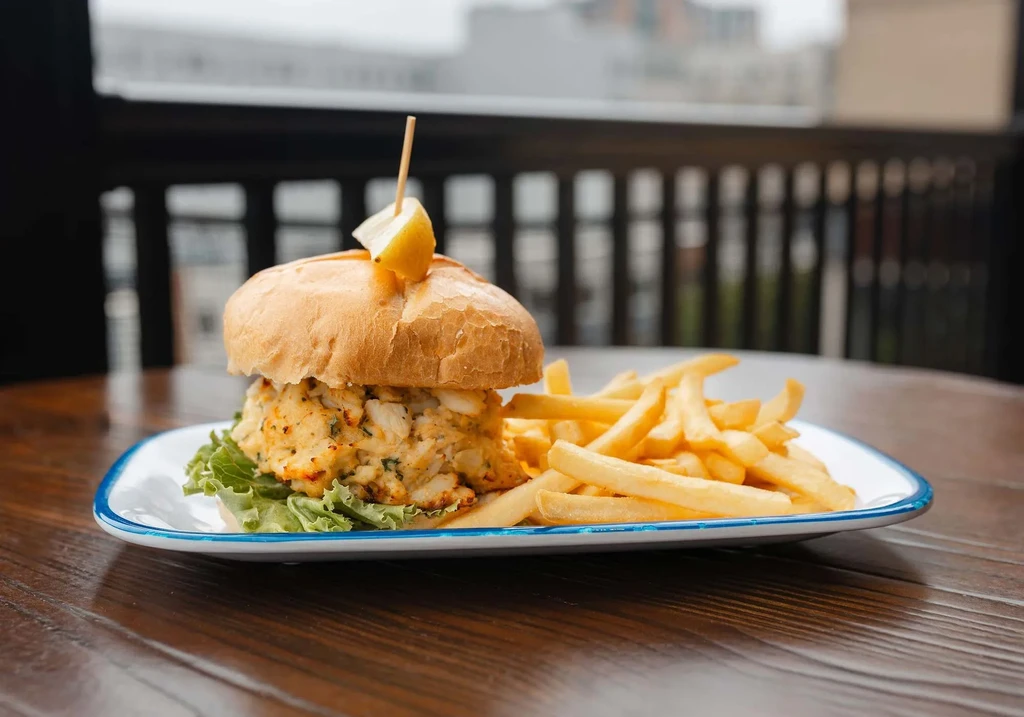 Crab cake sandwich, golden fries, and lemon on a plate outdoors.