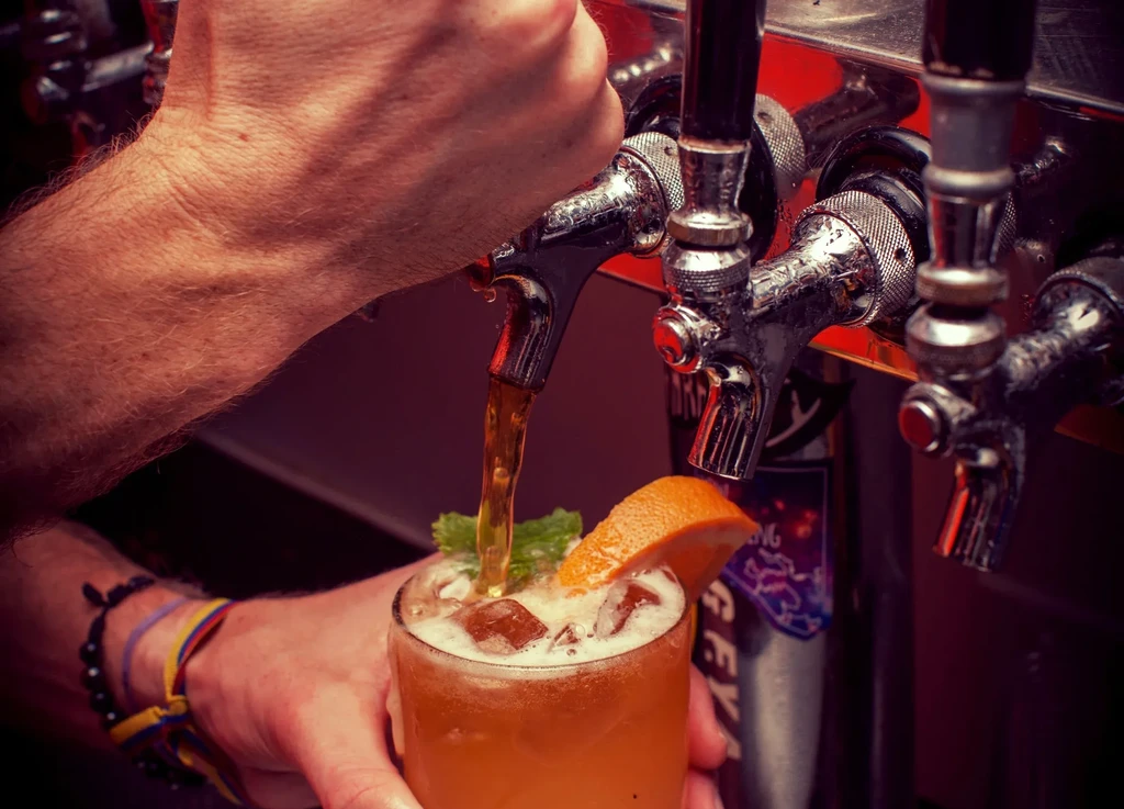 Hands pouring amber liquid from a tap into a garnished cocktail.