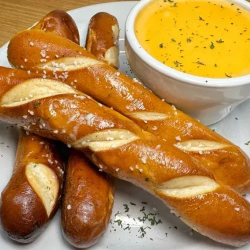 Golden-brown salted pretzel sticks and garnished yellow cheese dip on a plate.