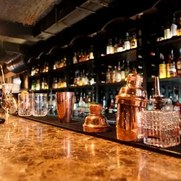 Marble bar counter with copper shakers, glassware, and backlit liquor bottles.