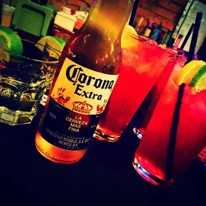 Corona Extra beer bottle and red cocktails on a dimly lit bar.