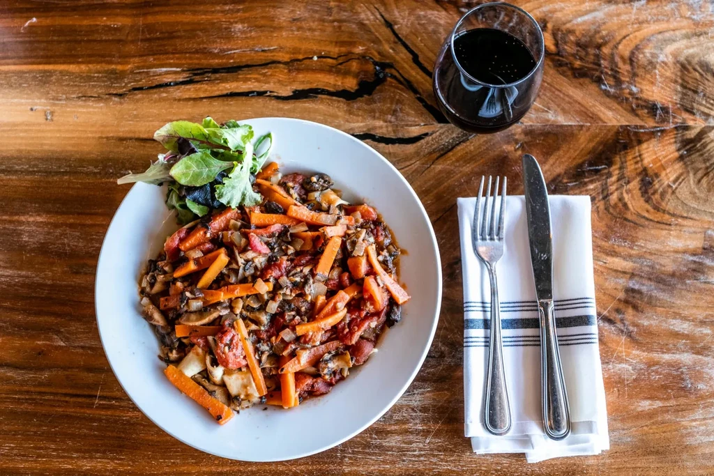 Colorful chopped vegetables, wine, and cutlery on a rustic wooden table.
